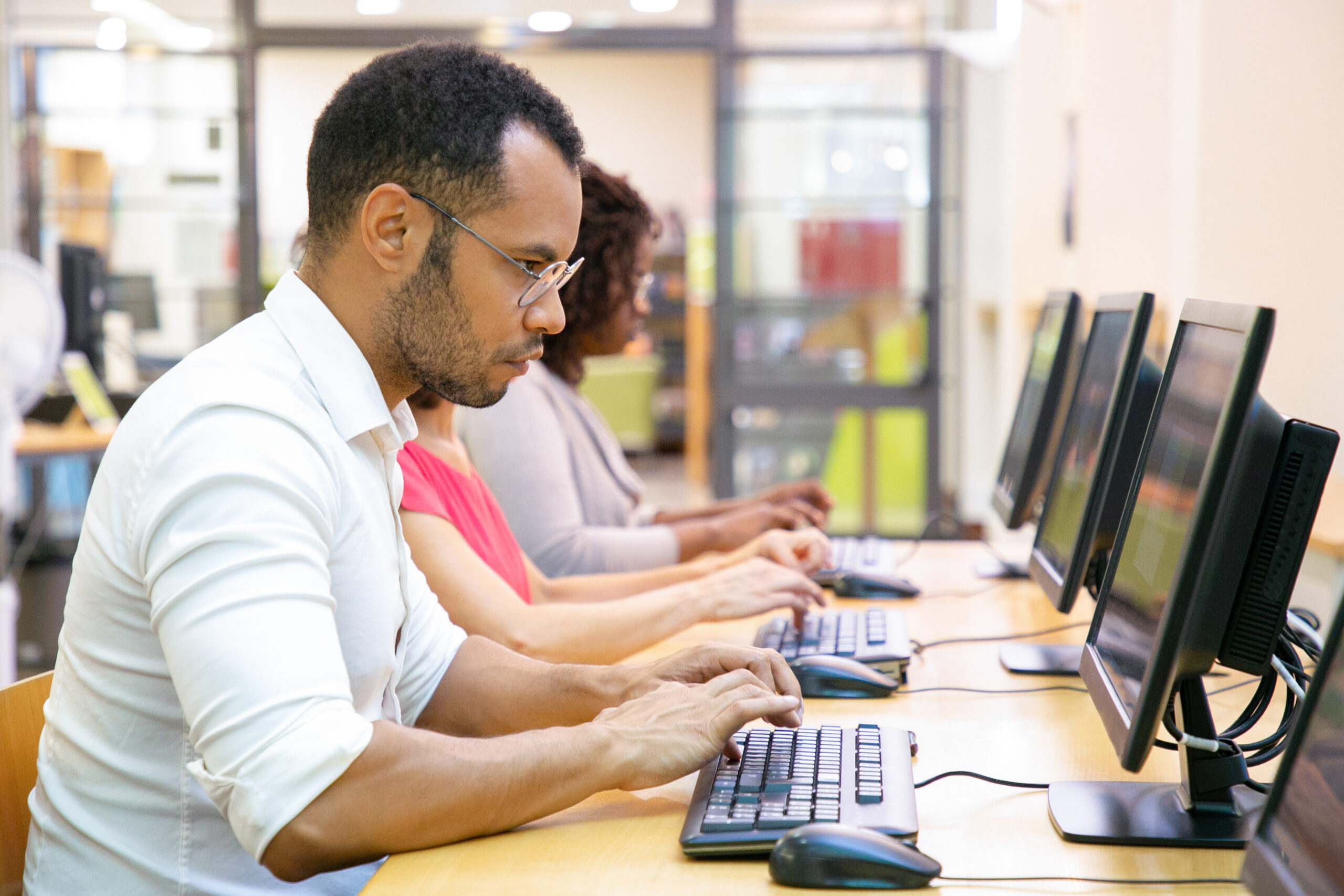 Extremely focused male student taking online test in computer class. Line of man and women in casual sitting at table, using desktops, typing, looking at monitor. Education center concept